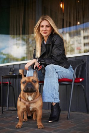 Photo for Beautiful blonde woman in a leather jacket sitting in a outdoor cafe with her French bulldog puppy on a leash. Attractive Ukrainian female person walking the dog outdoor - Royalty Free Image