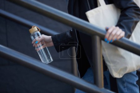 Foto de Persona femenina subiendo las escaleras con una botella de vidrio reutilizable con agua en la mano y una bolsa de algodón en el hombro. concepto de estilo de vida sostenible - Imagen libre de derechos