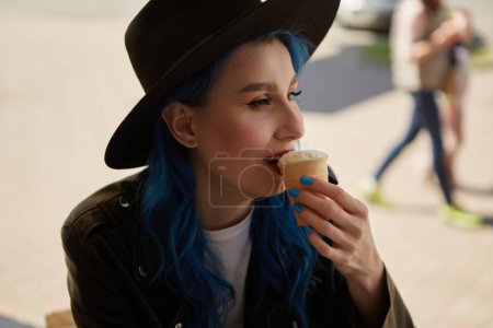 Foto de Retrato de una hermosa mujer diversa con el pelo teñido de azul comiendo un helado al aire libre. Atractiva mujer blanca con un elegante sombrero hipster y chaqueta de cuero disfrutando del postre en un café - Imagen libre de derechos