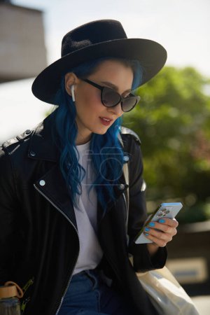Foto de Retrato de una mujer diversa con el pelo teñido de azul con un elegante sombrero hipster, chaqueta de cuero y gafas de sol. Hermosa joven escuchando música en auriculares inalámbricos y usando un teléfono inteligente - Imagen libre de derechos