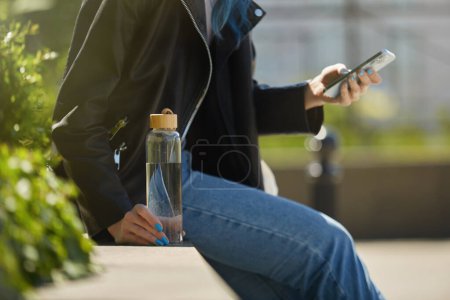 Téléchargez les photos : Jeune femme assise dans un jardin urbain sur le toit avec une bouteille d'eau douce en verre et à l'aide d'un smartphone. Femme méconnaissable naviguant sur une application mobile sur un téléphone moderne en plein air - en image libre de droit