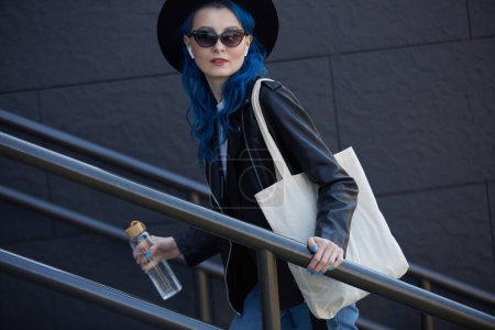 Téléchargez les photos : Modèle féminin diversifié avec cheveux bleus teints et sac fourre-tout sur l'épaule montant les escaliers dans la rue de la ville avec une bouteille d'eau en verre à la main. Concept de diversité et de durabilité - en image libre de droit