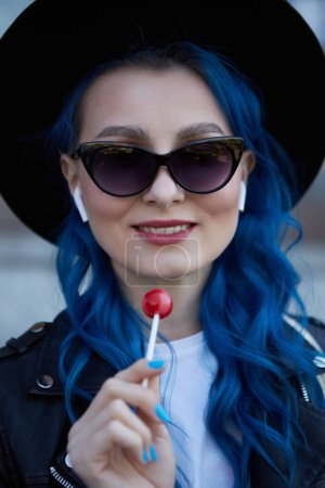 Foto de Retrato de una hermosa mujer diversa con el pelo teñido de azul lamiendo una piruleta. Mujer alegre que lleva un sombrero hipster, gafas de sol y escucha música en auriculares inalámbricos - Imagen libre de derechos