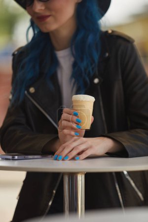Téléchargez les photos : Jeune femme diversifiée aux cheveux bleus teints mangeant et crème glacée en plein air. Femme en veste en cuir noir profitant d'un dessert sucré dans un café de rue - en image libre de droit