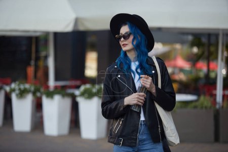 Foto de Hermosa joven con el pelo teñido de azul sosteniendo una botella de agua de vidrio en las manos. Diversa modelo femenina de pie en la calle de la ciudad con un café en el fondo - Imagen libre de derechos