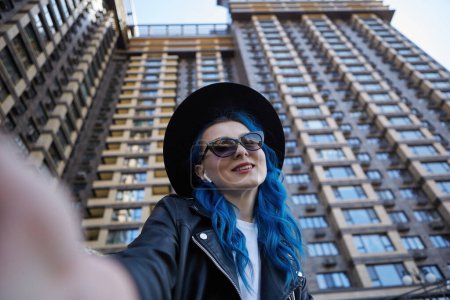 Photo for Diverse young woman taking a selfie. Beautiful white female with dyed blue hair takes a picture near high rise building in the city street - Royalty Free Image