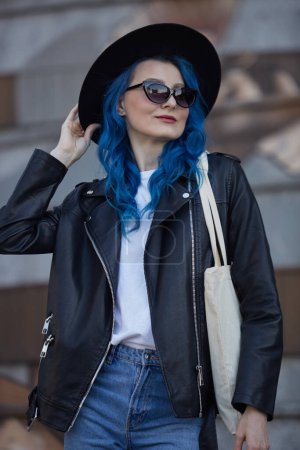 Téléchargez les photos : Belle femme diversifiée aux cheveux bleus. Jeune femme adulte portant un chapeau hipster élégant, une veste en cuir noir et un sac fourre-tout en coton dans la rue de la ville. - en image libre de droit