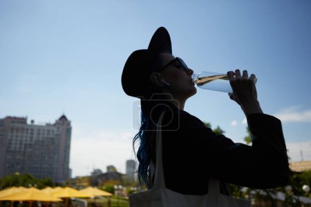 Foto de Silueta de mujer joven bebiendo agua de una botella de vidrio en la calle de la ciudad. Elegante persona femenina con el pelo teñido de azul con un sombrero hipster y bolso de mano en el hombro - Imagen libre de derechos