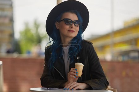 Foto de Retrato de una hermosa mujer de pelo azul en gafas de sol y sombrero hipster comiendo un helado en un café de la calle. Alegre persona femenina diversa disfrutando de un postre al aire libre en un día soleado - Imagen libre de derechos