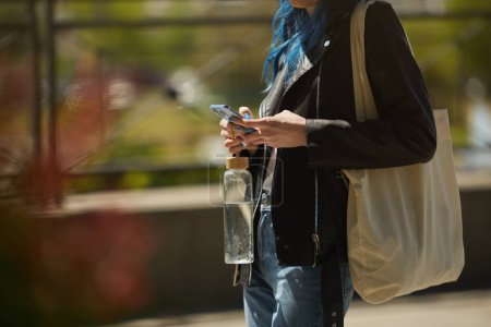 Foto de Mujer joven con bolsa de algodón en el hombro y sosteniendo una botella de agua de vidrio en la mano en un paseo. Persona femenina irreconocible usando un teléfono inteligente al aire libre - Imagen libre de derechos