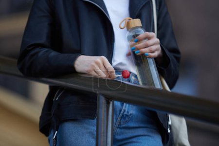 Foto de Mujer joven sosteniendo botella de agua de vidrio y caramelos de piruleta en la mano. Persona femenina irreconocible con chaqueta de cuero negro y vaqueros azules clásicos en un paseo - Imagen libre de derechos
