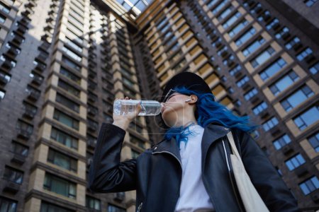 Foto de Persona femenina diversa que bebe agua fresca de una botella de vidrio cerca del edificio de gran altura en el centro de la ciudad. Hermosa mujer joven con pelo azul bebe líquido al aire libre - Imagen libre de derechos