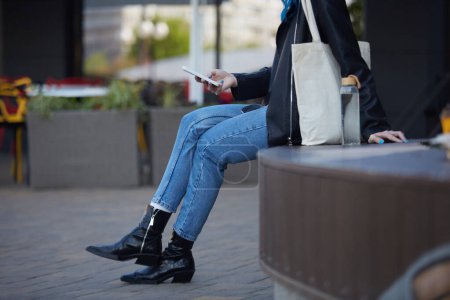 Foto de Mujer joven sentada en un banco con una botella de agua de cristal y la aplicación móvil de navegación en un teléfono inteligente. Persona femenina irreconocible que lleva un bolso de algodón en el hombro - Imagen libre de derechos