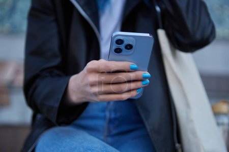 Téléchargez les photos : Jeune femme utilisant un smartphone bleu moderne avec triple caméra. Femme méconnaissable naviguant sur une application mobile sur un téléphone portable moderne en plein air - en image libre de droit