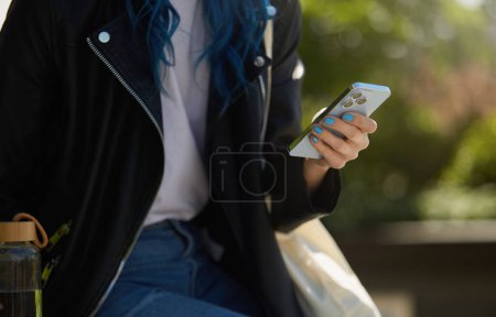Téléchargez les photos : Femme utilisant une application mobile sur un smartphone. Jeune femme méconnaissable en veste en cuir naviguant sur un réseau social sur un téléphone portable moderne - en image libre de droit