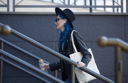 Foto de Diversa modelo femenina con el pelo teñido de azul caminando por las escaleras en el centro de la ciudad con una bolsa de algodón en el hombro y sosteniendo una botella de agua de vidrio en la mano - Imagen libre de derechos