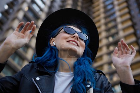 Photo for Portrait of a happy diverse woman enjoying favorite musical tracks. Beautiful white female with dyed blue hair listening to music in wireless headphones outdoor - Royalty Free Image