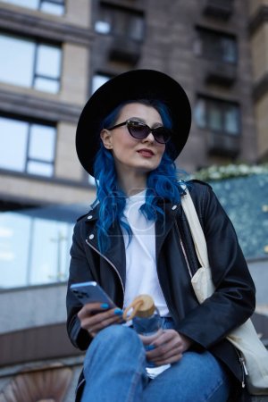 Foto de Hermosa persona diversa con el pelo azul sentado junto al edificio de gran altura al aire libre y sosteniendo un teléfono inteligente moderno y botella de agua de vidrio en las manos - Imagen libre de derechos