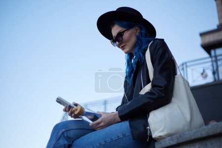 Foto de Elegante mujer diversa con una bolsa de mano y sombrero hipster al aire libre. Retrato de una hermosa hembra blanca sosteniendo una botella de vidrio de agua y un teléfono móvil moderno en las manos - Imagen libre de derechos