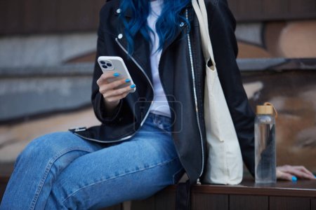 Téléchargez les photos : Jeune femme assise en plein air avec une bouteille d'eau en verre réutilisable et un sac à provisions en coton sur épaules et à l'aide d'un téléphone intelligent moderne - en image libre de droit