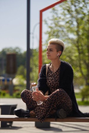 Téléchargez les photos : Jeune femme à la mode avec les cheveux courts teints assis sur un banc au soleil avec une bouteille d'eau en verre dans les mains. Modèle féminin diversifié en vêtements léopard relaxant au soleil en plein air - en image libre de droit