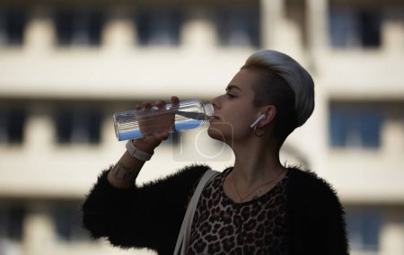 Téléchargez les photos : Jeune femme diversifiée boire de l'eau à partir d'une bouteille en verre extérieur. Femme élégante avec des boissons à cheveux teints courts liquide d'une bouteille réutilisable - en image libre de droit