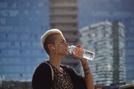 Foto de Retrato de una hermosa mujer de pelo corto bebiendo agua de una botella de vidrio al aire libre. Elegante joven mujer escuchando música en auriculares inalámbricos - Imagen libre de derechos