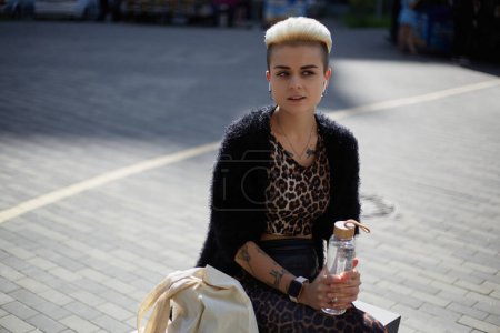 Foto de Hermosa hembra diversa con cabello corto y tatuajes sentados en un banco en el centro de la ciudad con una botella de agua de vidrio en las manos y escuchando música en auriculares inalámbricos - Imagen libre de derechos