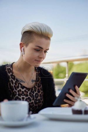 Téléchargez les photos : Belle jeune femme aux cheveux courts assise dans un café extérieur le matin, buvant du café et lisant un ebook sur tablette. Diverse personne féminine naviguant sur Internet sur un gadget - en image libre de droit