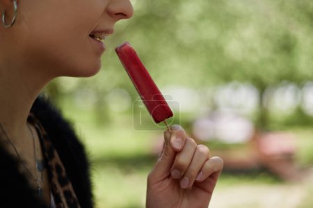 Téléchargez les photos : Jeune femme dégustant une glace aux canneberges sur bâton dans un parc verdoyant ensoleillé. Femme méconnaissable mangeant des desserts surgelés - en image libre de droit