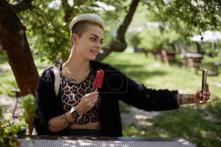 Photo for Happy diverse woman with short hair taking a selfie with an ice cream in a cozy outdoor cafe. Cheerful tom boy female posing for a selfie photo with a dessert in hand - Royalty Free Image
