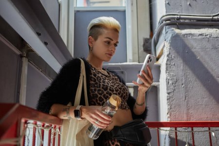 Foto de Mujer tatuada diversa con el pelo corto usando un teléfono inteligente con una botella de vidrio de agua en la mano y bolsa de mano en el hombro. Concepto de sostenibilidad y diversidad - Imagen libre de derechos
