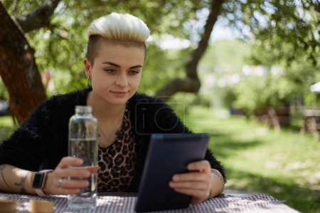 Téléchargez les photos : Belle jeune femme buvant de l'eau d'une bouteille en verre et lisant ebook sur tablette dans un parc verdoyant. Portrait de Tom Boy élégant femelle s'hydrater avec de l'eau douce - en image libre de droit