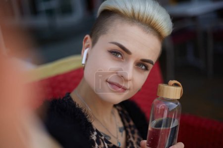 Photo for Beautiful diverse female with short hair posing for a selfie with a glass water bottle. Attractive white woman with tom boy hairstyle taking a selfie photo in a restaurant - Royalty Free Image