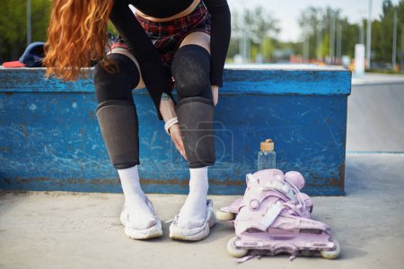Photo for Young skater girl putting on protective shin guards and knee pads for skating. Unrecognizable female roller blader preparing for a ride in a concrete skatepark in summer - Royalty Free Image
