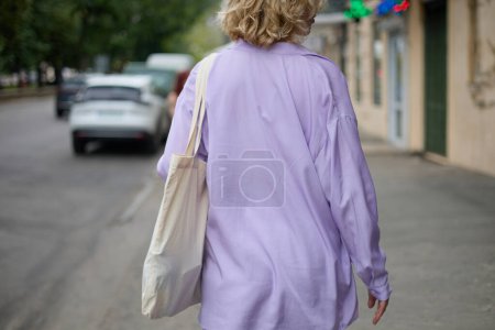 Téléchargez les photos : Jeune femme à la mode en vêtements décontractés violets marchant dans la rue avec un sac fourre-tout en coton sur l'épaule. Mode et concept de durabilité - en image libre de droit