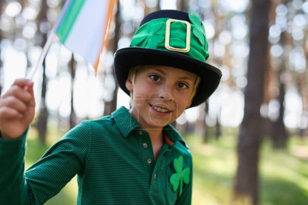 Glücklicher kleiner Kobold feiert den St. Patricks Day. Porträt eines niedlichen weißen Kindes, das mit der Flagge Irlands in grün schwenkt
