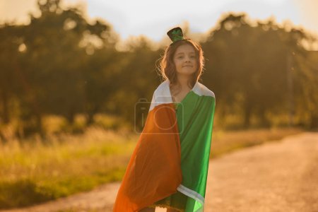 Photo for Adorable leprechaun girl covered with large Irish flag on a Saint Patrick's Day celebration - Royalty Free Image