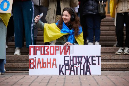 Téléchargez les photos : Joyeux patriote ukrainien prenant un selfie avec un drapeau national et une bannière "Budget transparent - Pays libre" lors d'une manifestation. Kiev - 23 mars 2024 - en image libre de droit