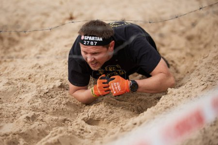 Foto de Hombre ucraniano arrastrándose sobre la arena bajo el alambre de púas durante la competencia de la Spartan Race en Kiev - 20 de abril de 2024 - Imagen libre de derechos