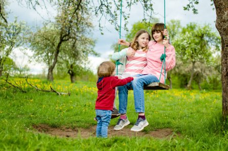 Photo for Two young sisters and their toddler brother having fun on a swing in blossoming apple orchard on warm spring day. Active outdoor activities for family with kids. - Royalty Free Image