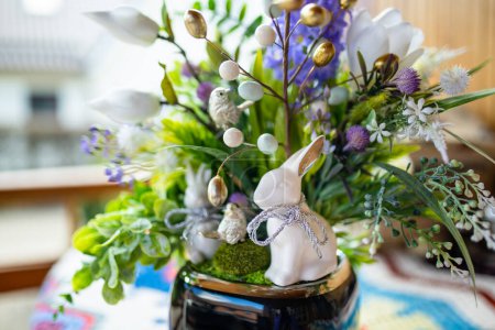 Photo for Easter table decoration made ot artificial flowers, green leaves, birds, and small eggs. White Easter bunny sitting in flower pot. - Royalty Free Image
