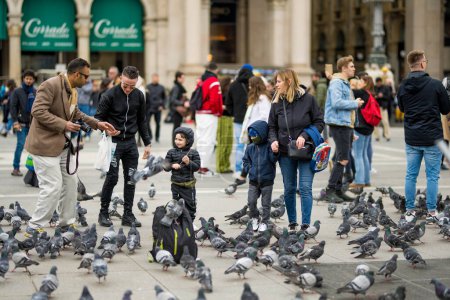 Foto de MILÁN, ITALIA - ABRIL 2022: Un grupo de personas alimentando a las palomas en la Plaza de la Catedral o Piazza del Duomo en el centro de Milán, Lombardía, Italia. - Imagen libre de derechos