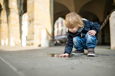 Photo for Cute toddler boy playing in a puddle on the street of Bergamo. Little child having fun exploring in Citta Alta, upper district of Bergamo. Bergamo, Lombardy, Italy. - Royalty Free Image