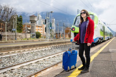 Téléchargez les photos : Une jeune femme dans une gare. Une fille attend un train sur un quai. Touriste féminine avec une valise prête à voyager. Partir en vacances seul. - en image libre de droit