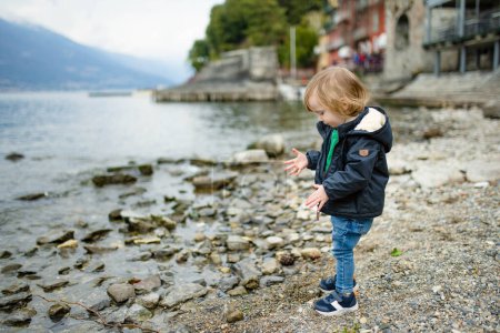 Téléchargez les photos : Petit garçon mignon jouant avec des cailloux dans le port de Varenna, l'une des villes les plus pittoresques sur la rive du lac de Côme. Varenna, Lombardie, Italie. - en image libre de droit