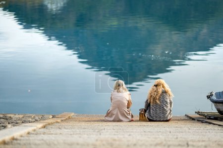 Photo for Two young women sitting at the embankment of Lecco town on early spring day. Friends enjoying picturesque waterfront of Lecco located on the Lake Como. Vacation destination in Italy. - Royalty Free Image