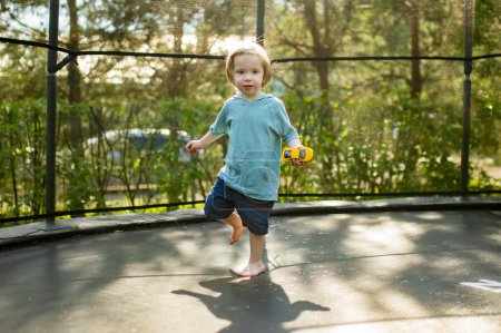 Foto de Lindo niño saltando en un trampolín en un patio trasero en un día de verano cálido y soleado. Deportes y ejercicios para niños. Actividades de ocio al aire libre de verano. - Imagen libre de derechos