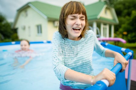Téléchargez les photos : Deux sœurs adolescentes drôles essayant d'entrer dans l'eau froide de la piscine. Les enfants s'amusent dans la piscine extérieure. Amusement en famille dans une piscine. Activités estivales pour la famille avec enfants. - en image libre de droit