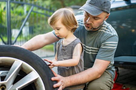 Netter kleiner Junge, der seinem Vater hilft, Autoräder im Hinterhof zu wechseln. Vater bringt seinem kleinen Sohn bei, Werkzeuge zu benutzen. Aktive Eltern eines kleinen Kindes.